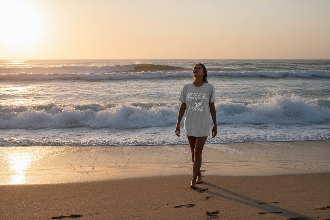 Person walking along an ocean shoreline at sunrise wearing a recovery graphic tee, representing sobriety and freedom